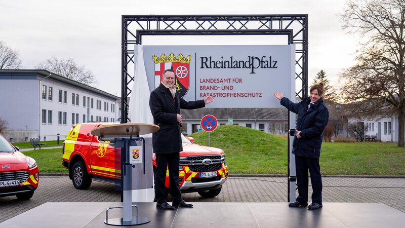 Innenminister Michael Ebling und Landesbrand- und Katastrophenschutzinspekteurin Kirstin Eisenhauer mit dem Schild der neuen Behörde. Im Hintergrund stehen landeseigene Einsatzfahrzeuge im ne