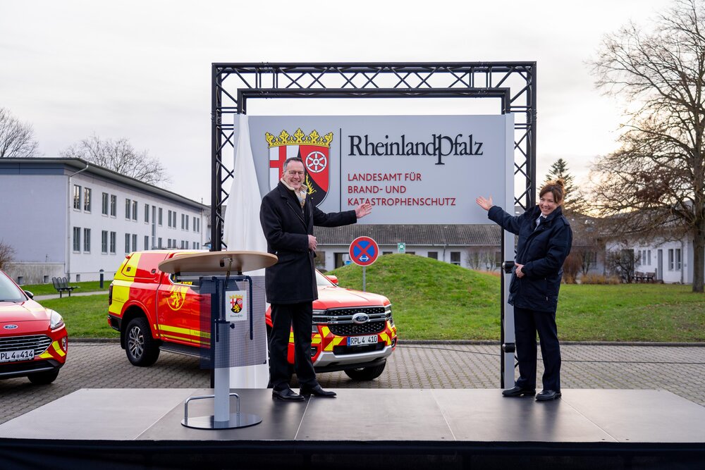 Innenminister Michael Ebling und Landesbrand- und Katastrophenschutzinspekteurin Kirstin Eisenhauer mit dem Schild der neuen Behörde. Im Hintergrund stehen landeseigene Einsatzfahrzeuge im ne